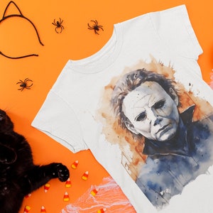 May include: A white t-shirt with a watercolor illustration of Michael Myers from the Halloween movie franchise. The shirt is surrounded by Halloween decorations, including a black cat, candy corn, and spiders.