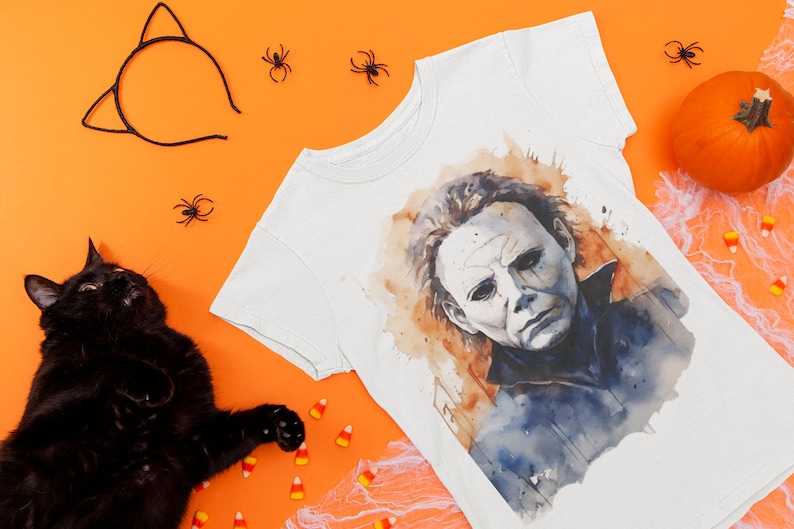 May include: A white t-shirt with a watercolor illustration of Michael Myers from the Halloween movie franchise. The shirt is surrounded by Halloween decorations, including a black cat, candy corn, and spiders.