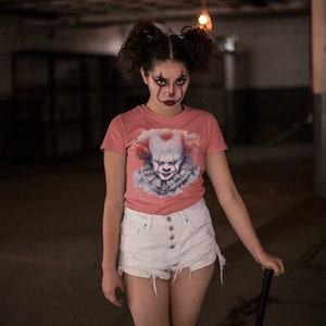 May include: A woman wearing a pink t-shirt with a graphic of a clown's face. She is also wearing white denim shorts and black stockings. She is holding a black and white baseball bat.