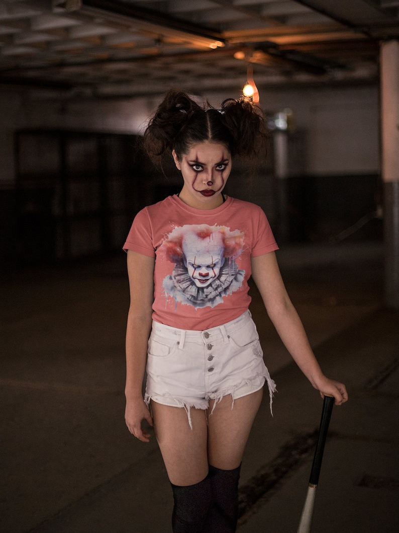 May include: A woman wearing a pink t-shirt with a graphic of a clown's face. She is also wearing white denim shorts and black stockings. She is holding a black and white baseball bat.