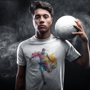 May include: A white t-shirt with a colorful graphic of a soccer player dribbling the ball. The graphic is painted in a watercolor style with splashes of color.