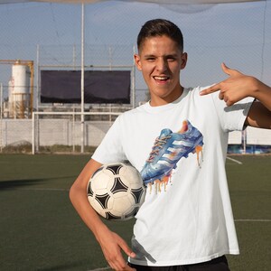 May include: A young person wearing a white t-shirt with a blue and orange watercolor design of a soccer cleat. The person is holding a soccer ball and pointing to the design on the shirt.