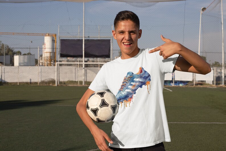May include: A young person wearing a white t-shirt with a blue and orange watercolor design of a soccer cleat. The person is holding a soccer ball and pointing to the design on the shirt.