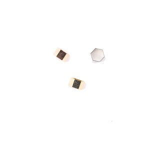 May include: Three pieces of jewelry on a white background. Two gold-colored rings with a dark square inset and a silver-colored, hexagonal-shaped piece. The rings are rectangular with a flat top and bottom.