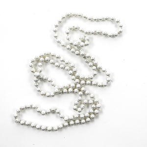 May include: A long strand of white pearl beads, strung together on a silver thread, is arranged in a loose, flowing pattern on a white background.