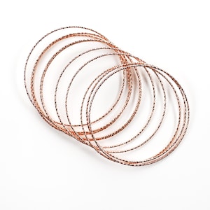 May include: A set of rose gold bangles, stacked and arranged in a circular pattern. The bangles are thin and have a textured surface.