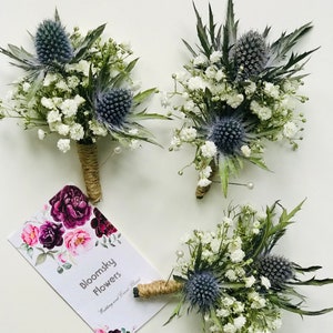 May include: Three floral buttonholes featuring blue thistle, white baby's breath, and green foliage, with stems wrapped in twine and pearl-tipped pins. A rectangular card displays the text Bloomsky Flowers Wedding and Event Florist.