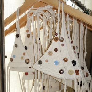May include: Cream-colored knitted triangle tops with various colored buttons, hanging on a wooden hanger. The tops have long, cream-colored ties and are displayed in a well-lit setting.
