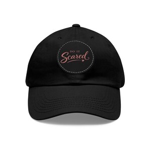 May include: Black baseball cap with a curved peak. A circular patch on the front displays the words "Do It Scared" in a cursive font, with a small heart. The text and heart are in a rose colour, set against a white dotted circle.