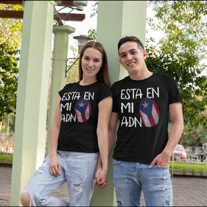 Op de afbeelding: Twee mensen dragen zwarte T-shirts met een witte print die "ESTA EN MI ADN" zegt en een ontwerp van de Puerto Ricaanse vlag. De vlag is een vingerafdruk in de kleuren rood, wit en blauw.