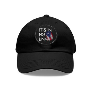 Op de afbeelding: Zwarte baseballpet met een witte geborduurde patch waarop "It's in my DNA" staat en een Puerto Ricaanse vlag.