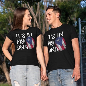 Puede incluir: Camisetas negras con el texto "IT'S IN MY DNA" y un diseño de la bandera de Puerto Rico. El diseño de la bandera está integrado en forma de huella dactilar. Las camisetas las lleva una pareja, de la mano, en un entorno exterior.