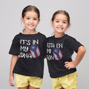 May include: Two children wearing black t-shirts with a Puerto Rican flag design. The text on the shirts reads "It's in my DNA" and "Esta en mi ADN".