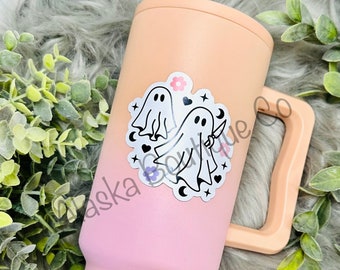 May include: A pink and peach ombre stainless steel tumbler with a ghost sticker featuring two ghosts, one holding a knife. The sticker also includes hearts, stars, and crescent moons. The tumbler has a handle and the word "slim" printed on the bottom.