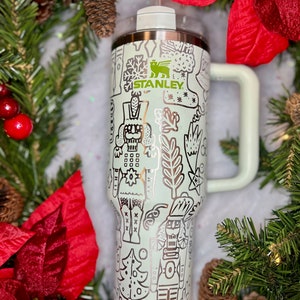 May include: A light green stainless steel tumbler with a white lid and a straw. The tumbler has a gold metallic design of a nutcracker, snowflakes, and other Christmas-themed illustrations. The tumbler has the word "STANLEY" printed on it.