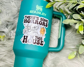 May include: A teal green Stanley travel mug with a black and white sticker that says "There's some horrors in this house". The sticker features a ghost, a mummy, and a pumpkin. The mug has a handle and a straw.