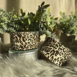 Op de afbeelding: Twee cilindrische containers met een luipaardprint. Eén container staat rechtop, de andere is gekanteld. De containers staan op een zilveren oppervlak, met een achtergrond van imitatiebont en groen in de achtergrond.