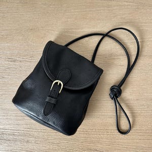 May include: Black leather crossbody bag with a flap closure and a gold-tone buckle. The bag features a long, adjustable strap and a textured surface. The bag is approximately 7 inches tall and 6 inches wide.