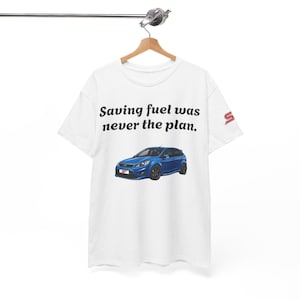 May include: A white short-sleeve t-shirt featuring the black text "Saving fuel was never the plan." above a graphic of a blue hatchback car. The sleeve displays a red and white "ST" logo. The garment is displayed on a wooden hanger.