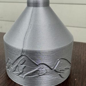 May include: A silver-colored, 3D-printed decorative vase with a mountain and leaf design around the base. The vase has a cylindrical base and a conical top with a rounded finial. The surface has a textured, ridged appearance.