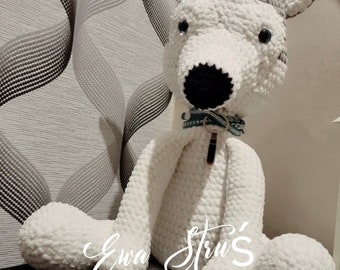 Puede incluir: Un perro de crochet blanco con ojos y nariz negros, que lleva un collar con estampado verde y blanco con una etiqueta plateada. El perro está sentado sobre una superficie blanca y negra con el texto "Ewa Stru's" visible.