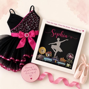 May include: A black dance leotard with a pink satin bow, a white shadow box frame containing a silhouette of a ballerina and various dance pins, a round pink box with the text "Dancing is not just a hobby, it's a way of life.", and pink ballet slippers.