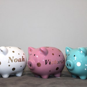 May include: Three ceramic piggy banks with gold polka dots. The piggy banks are white, pink, and teal. The white piggy bank has the name "Noah" written on it. The pink piggy bank has the name "Victoria" written on it. The teal piggy bank has the name "James" written on it.