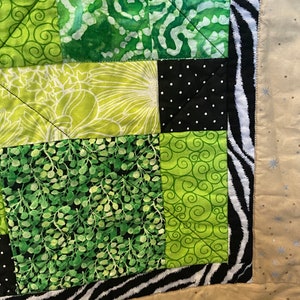 Puede incluir: Un edred&oacute;n hecho a mano con un dise&ntilde;o de patchwork. El edred&oacute;n presenta cuadrados de tela verde con varios patrones, incluyendo espirales, estampados florales y lunares. Un borde con estampado de cebra enmarca el edred&oacute;n, con un reverso beige.