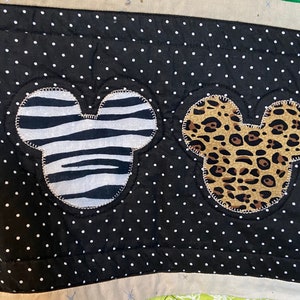 Puede incluir: Primer plano de una tela de lunares blancos y negros con dos formas de cabeza de Mickey Mouse. Una cabeza es con estampado de cebra, la otra con estampado de leopardo. La tela tiene un borde beige con detalles de estrellas plateadas.
