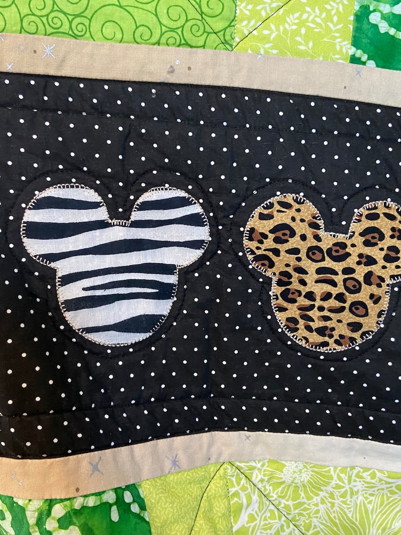 Puede incluir: Primer plano de una tela de lunares blancos y negros con dos formas de cabeza de Mickey Mouse. Una cabeza es con estampado de cebra, la otra con estampado de leopardo. La tela tiene un borde beige con detalles de estrellas plateadas.