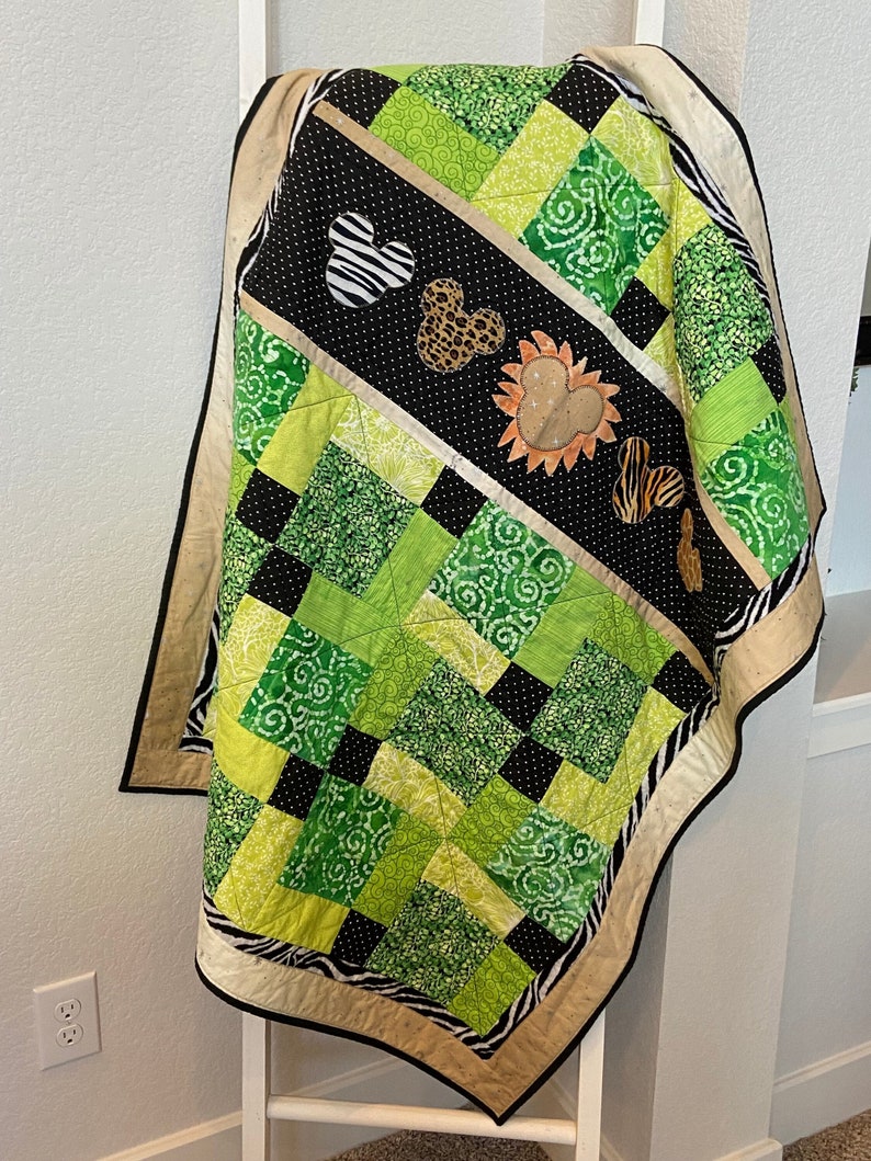 Puede incluir: Un edred&oacute;n hecho a mano con un dise&ntilde;o de patchwork en tonos verdes, negros y beige. El edred&oacute;n presenta detalles de estampado animal, incluyendo patrones de cebra y leopardo, y apliques con forma de Mickey Mouse. El edred&oacute;n est&aacute; colgado sobre una escalera blanca.