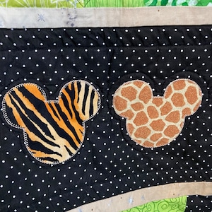 Puede incluir: Un edred&oacute;n colorido con formas de cabeza de Mickey Mouse con estampados de animales. Una cabeza tiene rayas de tigre y la otra manchas de jirafa. El fondo es negro con lunares blancos y tela verde estampada.