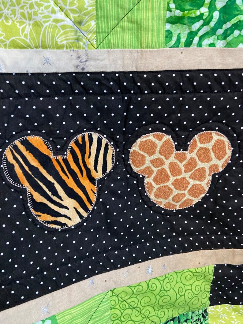 Puede incluir: Un edred&oacute;n colorido con formas de cabeza de Mickey Mouse con estampados de animales. Una cabeza tiene rayas de tigre y la otra manchas de jirafa. El fondo es negro con lunares blancos y tela verde estampada.