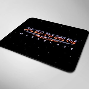 Op de afbeelding: Een zwarte muismat met een wit en oranje logo met de tekst "Xenon Megablast" op een zwarte achtergrond met witte sterren.