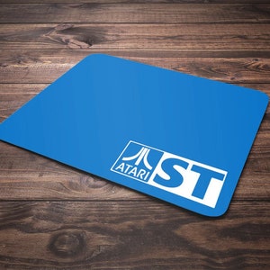 May include: A rectangular blue mouse pad featuring the white Atari logo and the letters ST in a white rectangular box located in the bottom right corner. The surface of the mouse pad is smooth and solid blue, providing a clean and simple design.