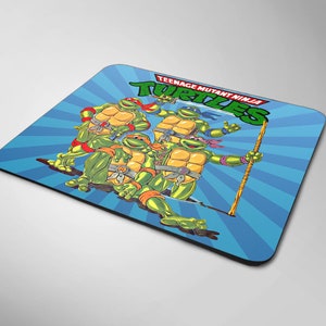 Puede incluir: Un ratón de ordenador azul y blanco con un gráfico de las Tortugas Ninja. Las tortugas son verdes con máscaras rojas y están en una pose de lucha. El texto "Teenage Mutant Ninja Turtles" está escrito en blanco sobre una pancarta roja.