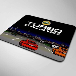 Puede incluir: Alfombrilla de ratón negra con una escena de videojuego retro con coches pixelados corriendo en una pista. La alfombrilla tiene el texto "TURBO CHALLENGE 2" y el logotipo de Lotus.