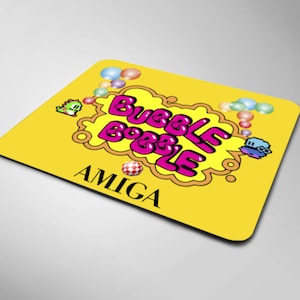 May include: Yellow mousepad with a pink "Bubble Bobble" logo and a small blue and white character. The logo is surrounded by a yellow cloud with white outlines. The mousepad also has the word "AMIGA" in black text.