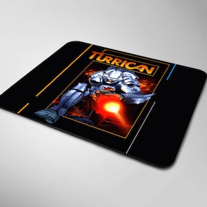 Pode incluir: Um tapete de rato retangular preto com o logótipo do videojogo Turrican em texto laranja no topo. O gráfico central mostra uma personagem com armadura prateada a disparar um raio de energia vermelho brilhante contra um fundo laranja e preto.