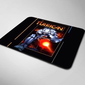 May include: A black mouse pad with a graphic of a futuristic robot firing a weapon. The robot is silver and the background is a fiery orange and yellow. The text "TURRICAN" is in yellow letters.