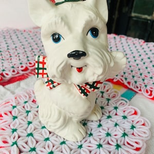 May include: A white ceramic Scottie dog figurine with blue eyes, a black nose, and a red tongue. The dog wears a plaid bow tie and a hat with a red pom-pom. The figurine is sitting on a white and red crocheted doily.