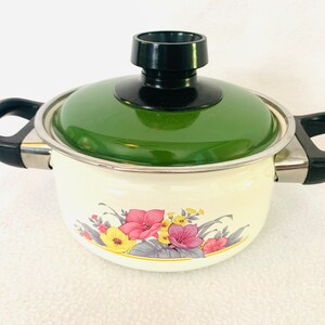 May include: A white enamel saucepan with a green lid and black handles. The saucepan has a floral design with pink, yellow, and purple flowers.