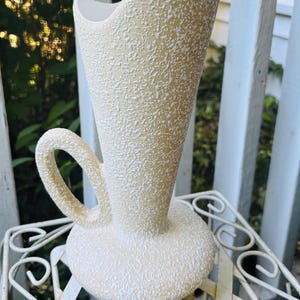 May include: Cream-colored ceramic vase with a textured surface and a handle. The vase has a wide, open top and a rounded base. The handle is curved and attached to the side of the vase. The vase is sitting on a white metal table.