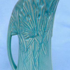 May include: A turquoise ceramic pitcher with a curved handle and a wide, stylized spout. The pitcher features a raised, textured design of leaves and lines, and has a crackled glaze finish. The pitcher is a decorative item.