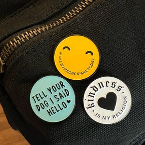 May include: Three enamel pins on a black fabric background. The first pin is yellow with a smiley face and the text "Make someone smile today". The second pin is light blue with the text "Tell your dog I said hello" and a small heart. The third pin is white with the text "Kindness is my religion" and a black heart.