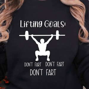 Puede incluir: Sudadera negra con texto blanco que dice "Lifting Goals: Don't fart Don't fart Don't fart" y una silueta de una persona levantando pesas.