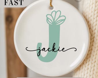 May include: A white ceramic ornament with a gold string for hanging. The ornament features a large, light teal letter "J" with a floral design and the name "jackie" in a cursive font. The text "MAKE IT CUSTOM" is at the bottom.