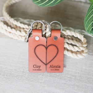 May include: Two matching leather keychains in a warm orange hue, each with a half-heart design that forms a complete heart when placed together. The names "Clay" and "Alexia" are engraved, along with the date "7.17.2021".