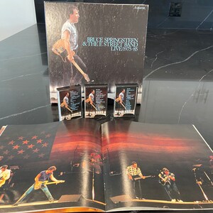 Puede incluir: Un álbum de vinilo negro con una foto de Bruce Springsteen en la portada. El título del álbum es "Bruce Springsteen & The E Street Band Live 1975-85". Tres cintas de cassette están delante del álbum. El álbum está abierto en una página con una foto de Bruce Springsteen y su banda actuando en el escenario.