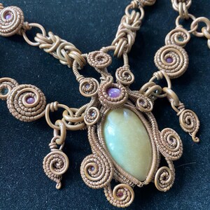 May include: A gold wire wrapped necklace with a large green stone pendant and smaller purple stones. The necklace is made of multiple strands of wire that are woven together.